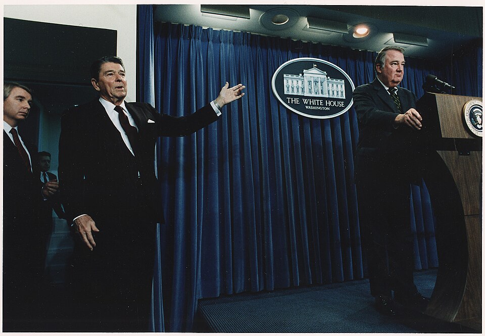US Attorney General Edward Meese stands at the podium in the White House Press Briefing Room addressing the Iran Contra Scandal, President Reagan stands far to the side and points his right hand at Reese.