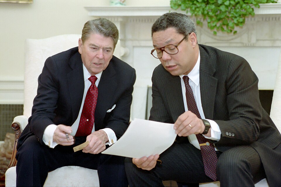President Reagan and National Security Advisor Colin Powell seated in the Oval Office and having a discussion.