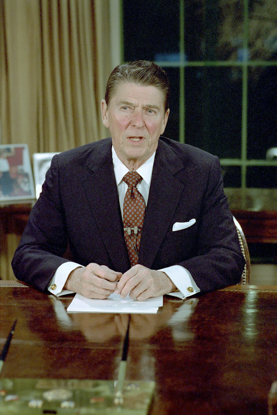 President Ronald Reagan in the Oval Office, seated at the resolute desk, giving an address to the nation.